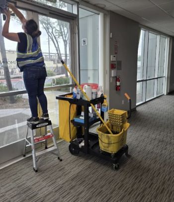 Janitorial worker on step ladder cleaning high windows in an office hallway beside a cleaning cart